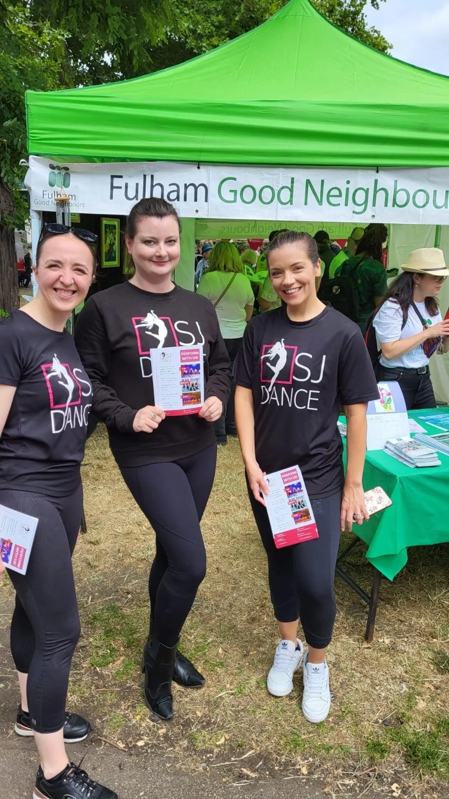 Three women wearing black dancer T-shirts standing in front of a Fulham Good Neighbours tent at an outdoor community event, smiling and holding brochures.