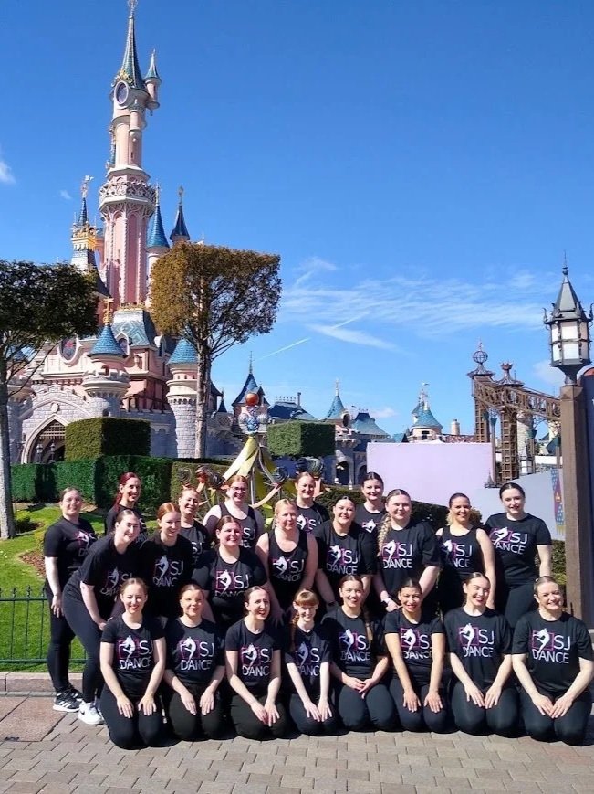 A group of dancers posing in front of Sleeping Beauty Castle at Disneyland, with colourful spires and a blue sky in the background.
