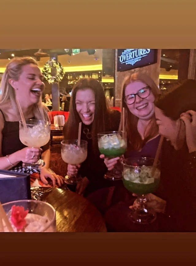 Four women sitting at a bar table, laughing and holding large cocktails with straws, in a lively indoor setting.