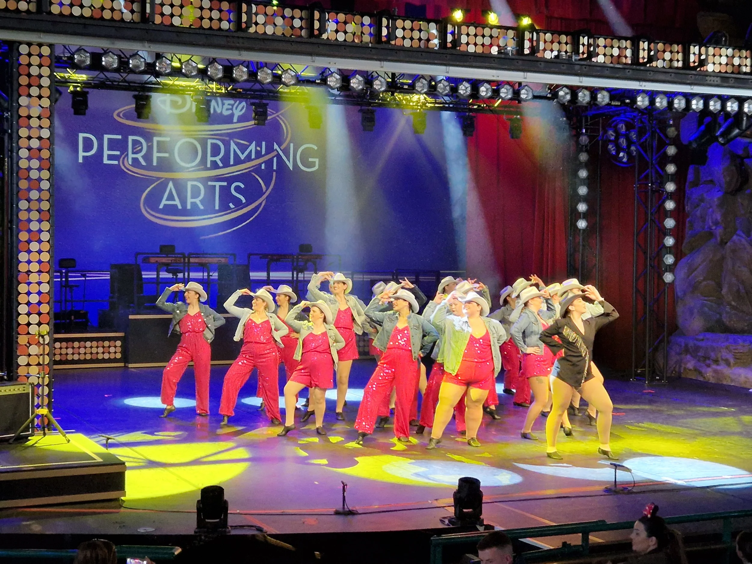 A group of performers dressed in cowboy-themed costumes with pink costumes, denim jackets, and silver hats dance on stage with a Disney Performing Arts sign in the background, illuminated by colourful stage lights.