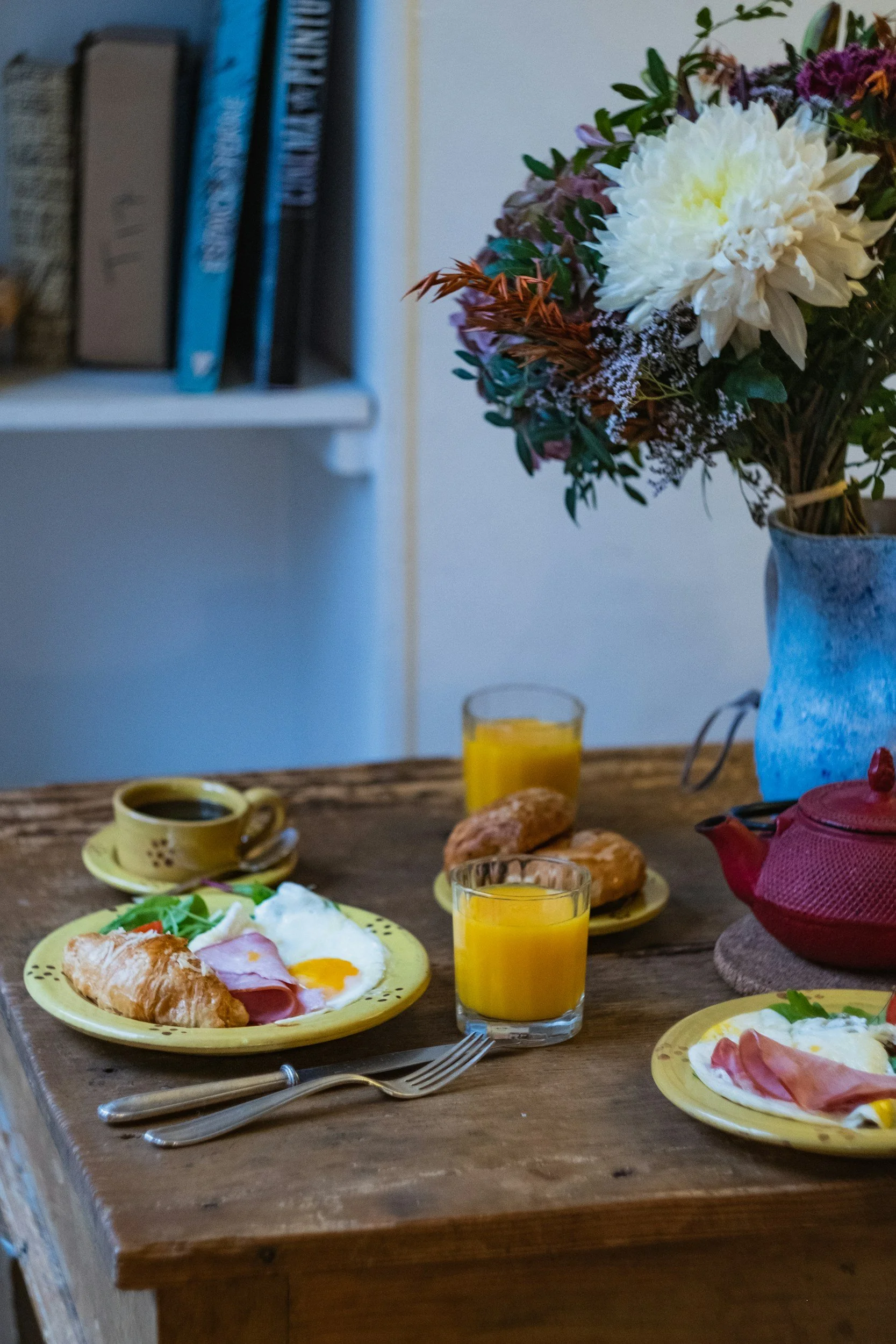 A breakfast table set with yellow plates and cups, containing eggs, ham, croissants, salad, and butter, with glasses of orange juice, on a wooden table. There is a flower arrangement in a blue vase and a bookshelf in the background.