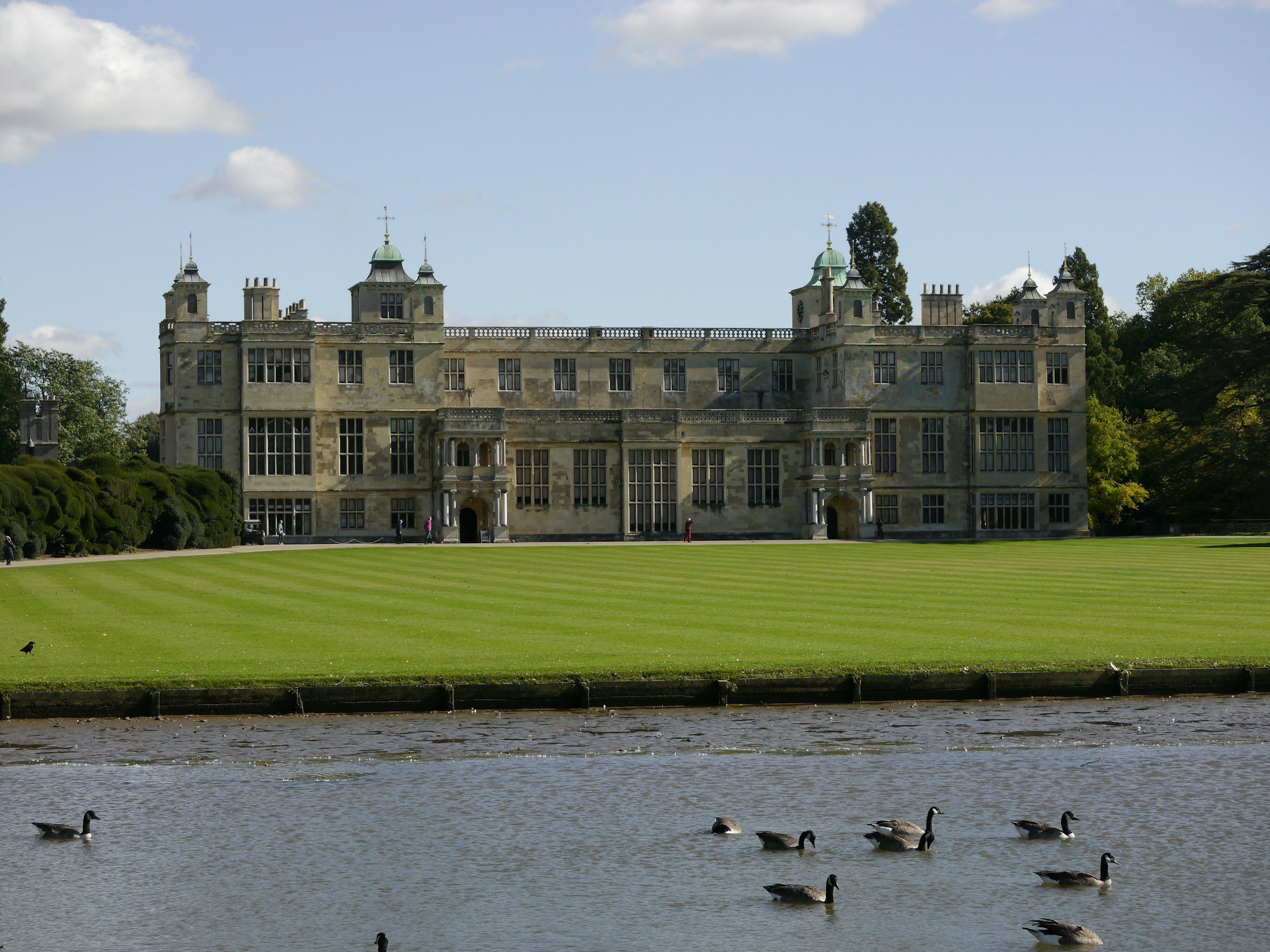 A large historic castle with towers and a green roof, surrounded by lush green trees, with a well-maintained lawn in front. Ducks are swimming in a pond in the foreground.