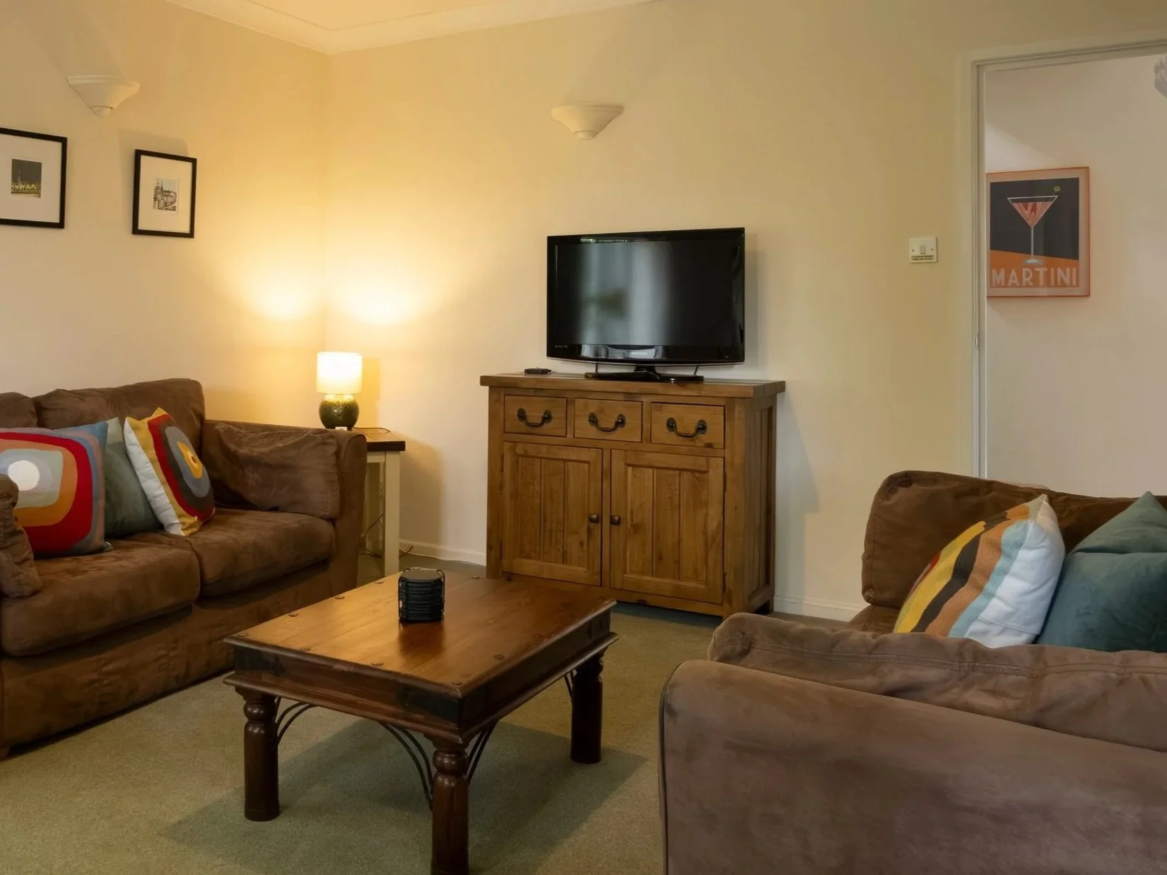 A cozy living room with beige walls, a brown sofa with colorful patterned pillows, a wooden coffee table, a TV on a wooden stand, and a side table with a lamp. There are framed pictures on the wall, a window with white curtains, and a ceiling light f