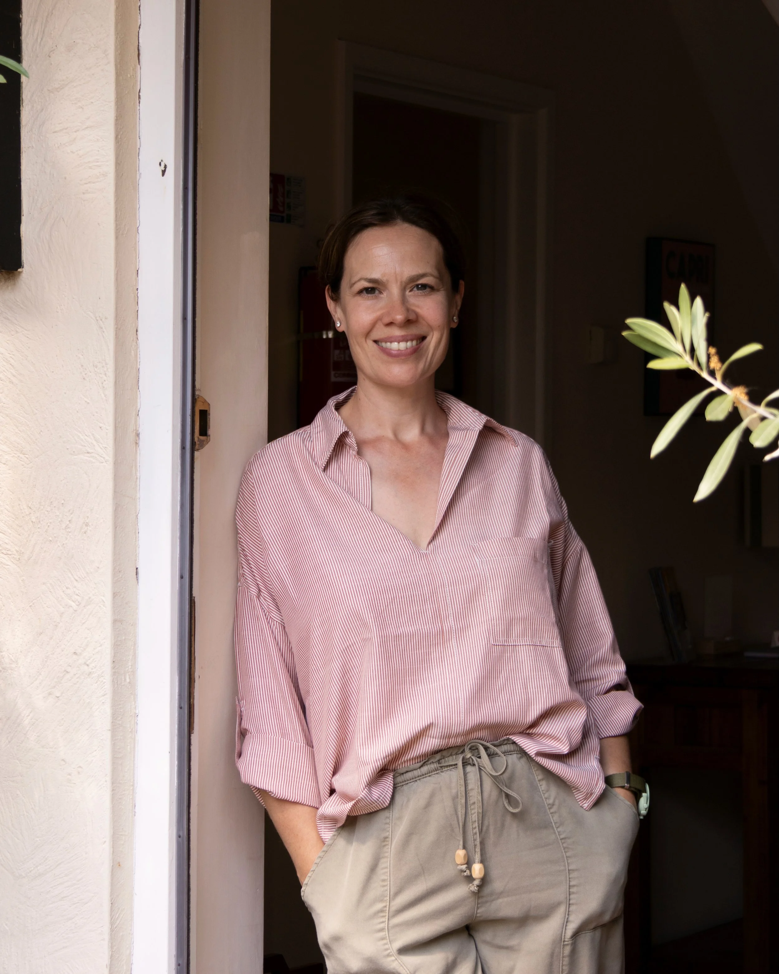 A woman with short brown hair, wearing a pink and white striped shirt and beige shorts, standing at a doorway with a smile.