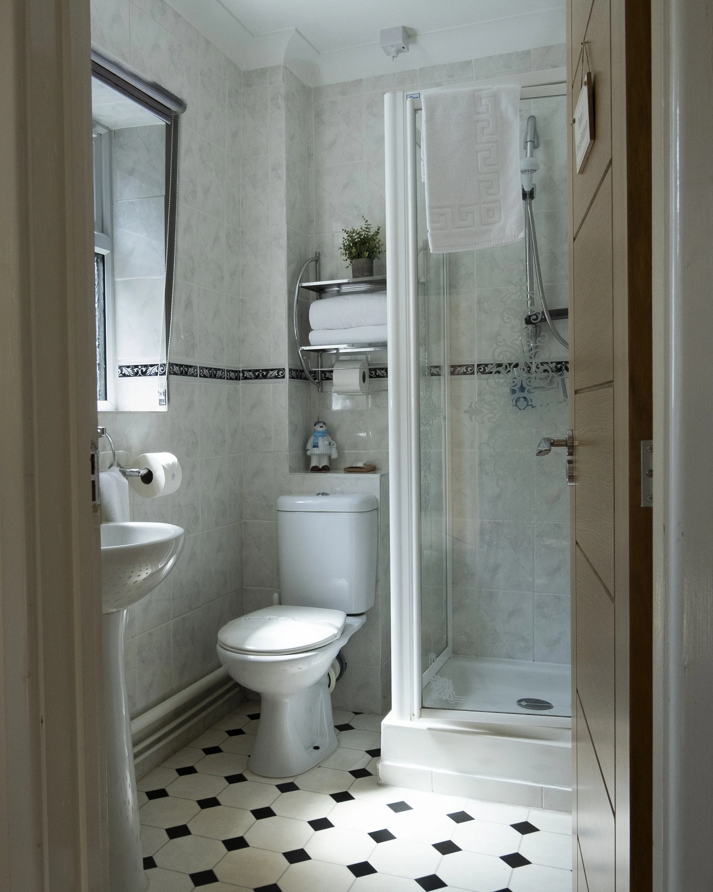 A small bathroom with a toilet, a sink, a glass shower enclosure, and a window. The walls and floor are tiled, and there is a decorative black and white tile border. A towel hangs on the shower door, and a small potted plant and towels are on a tray 