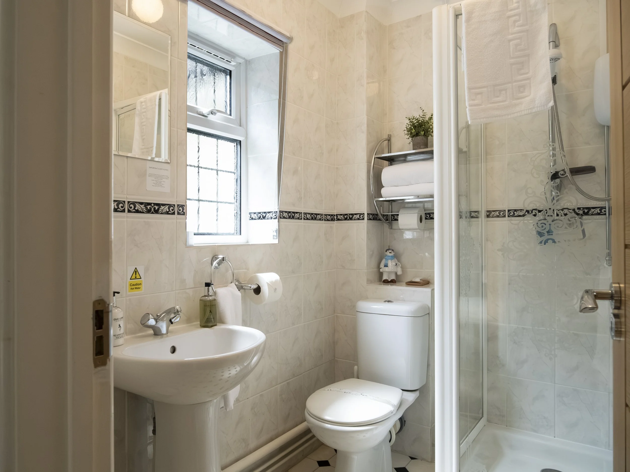 A small bathroom with beige marble tile walls, a frosted window, a white ceramic sink with two faucets, a toilet, a shower with a sliding glass door, a towel rack with a white towel, and a small potted plant on a shelf.