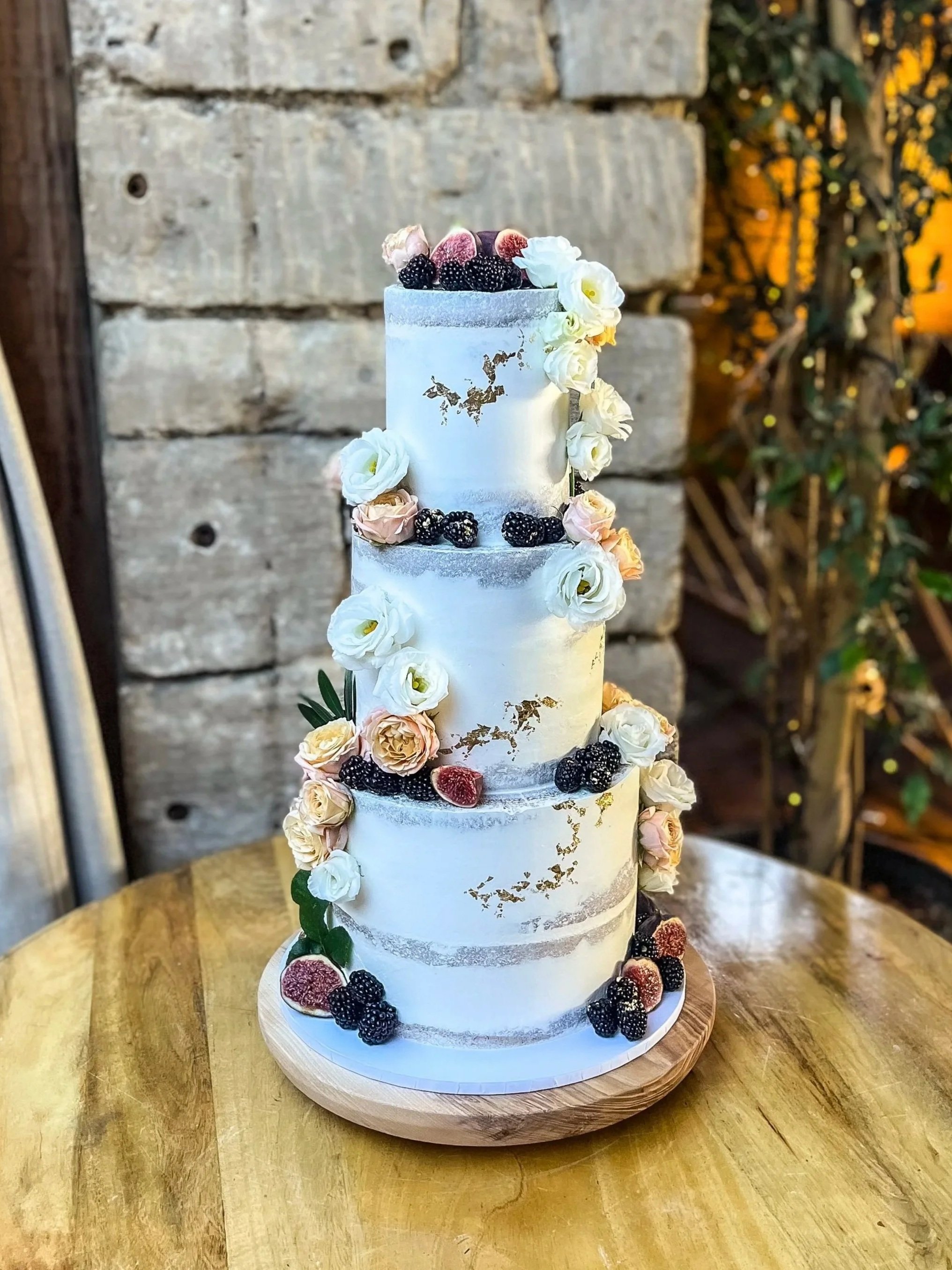 A tall, multi-tiered white cake decorated with blackberries, raspberries, and white flowers, placed on a wooden cake stand on a wooden table outdoors.