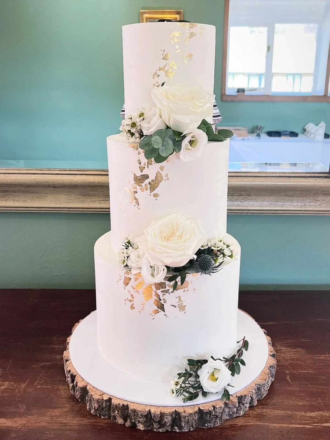A tall, three-tiered white cake with gold leaf accents, decorated with white roses, lisianthus, eucalyptus, and other greenery, sitting on a wooden slab.