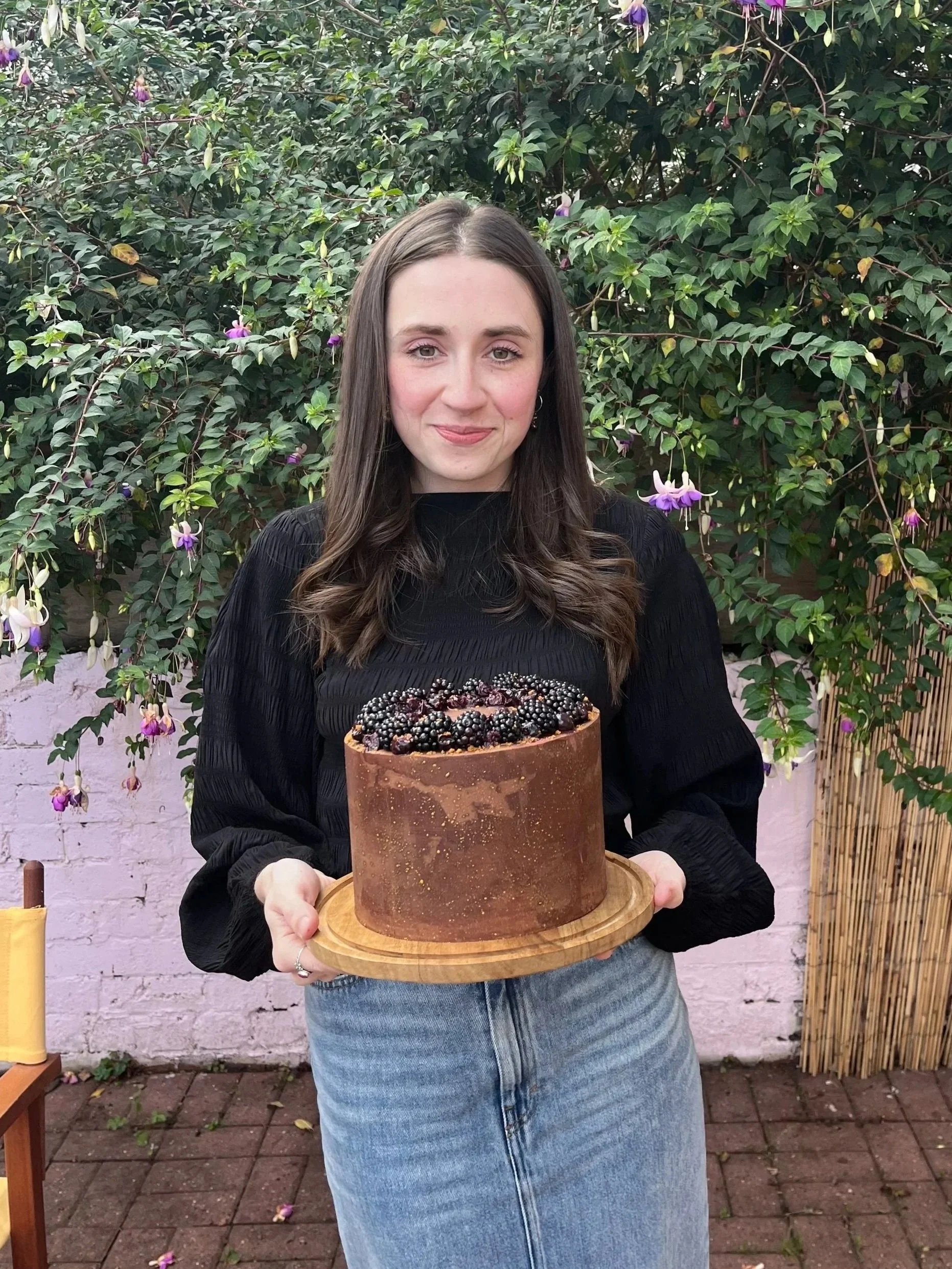 A woman with long brown hair and a black top holding a chocolate cake topped with blackberries and purple flowers in front of a green bush with purple and white flowers.