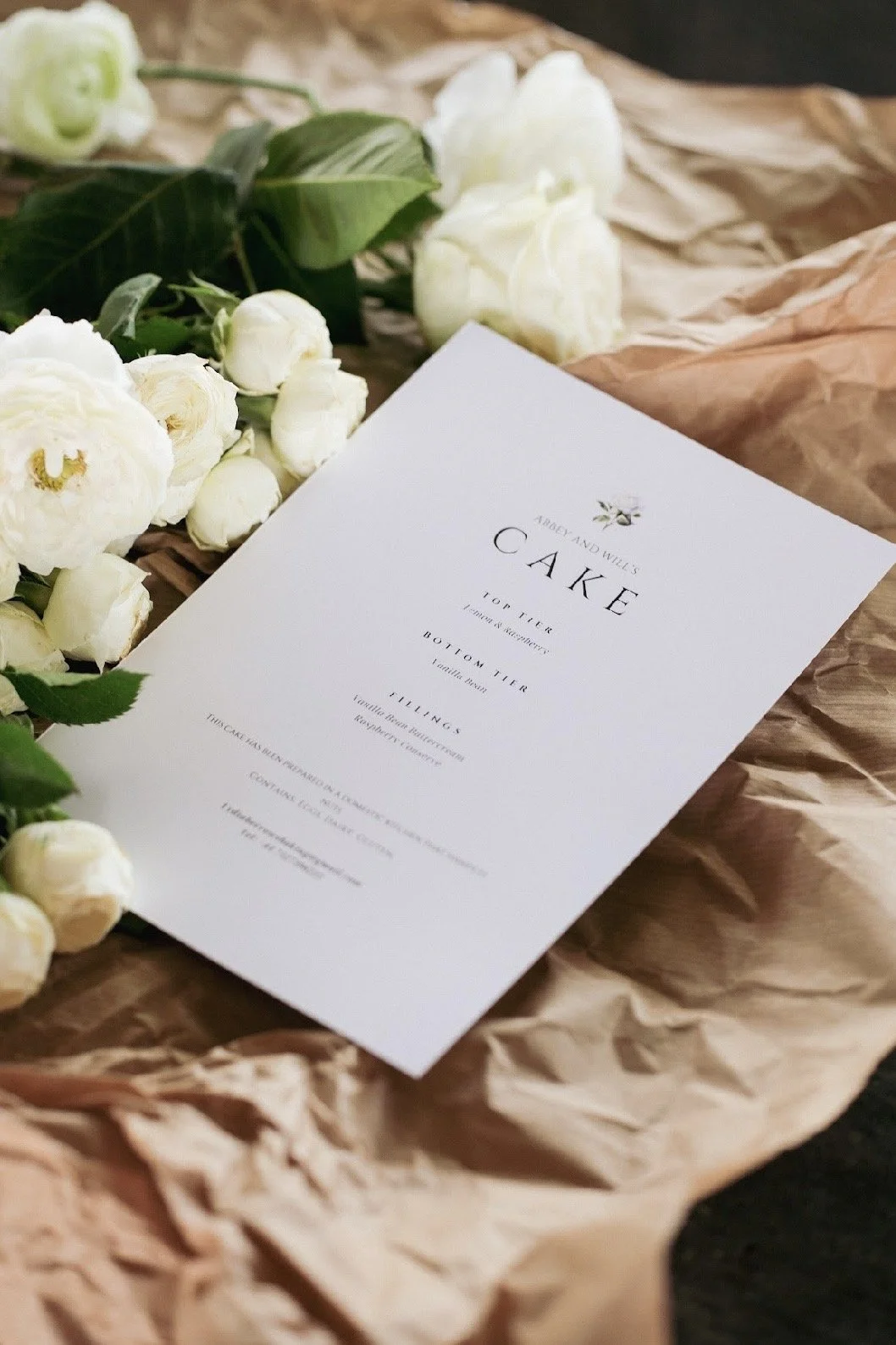 A wedding cake recipe sheet placed on crumpled brown paper, surrounded by white roses and green leaves.