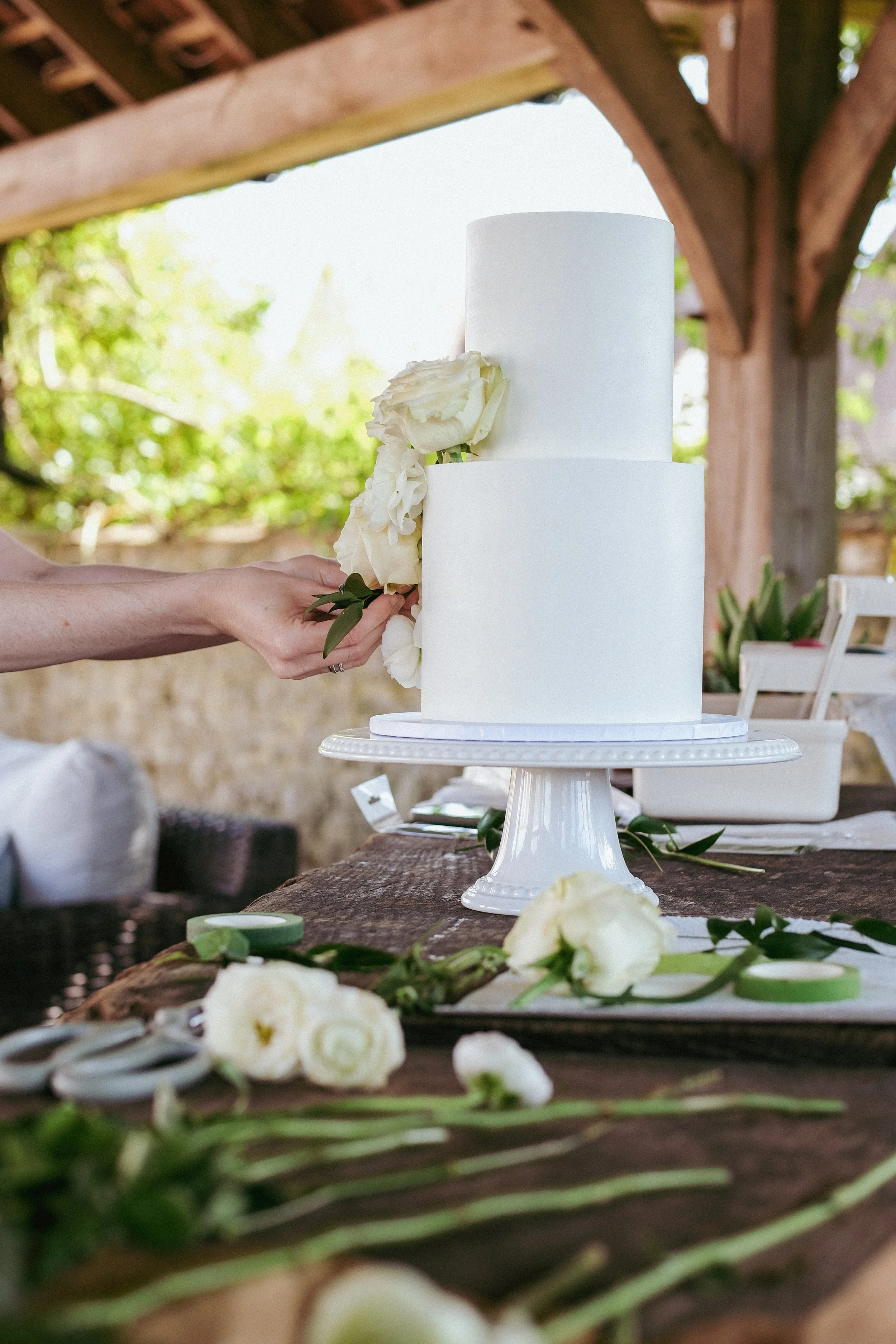 A person arranging white flowers on a white, tiered wedding cake outdoors, with a wooden structure and green foliage in the background.