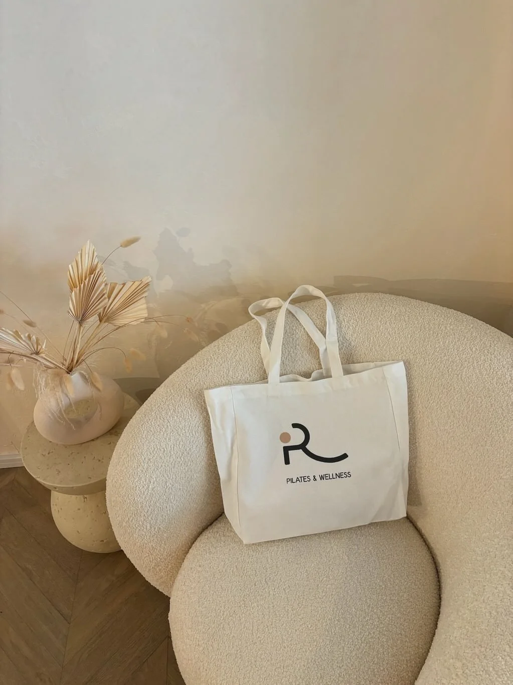 A white tote bag with a logo and the words 'Pilates & Wellness' sits on a round beige textured chair. Next to the chair is a small beige side table holding a vase with dried beige palm leaves. The background features a plain, light-colored wall.