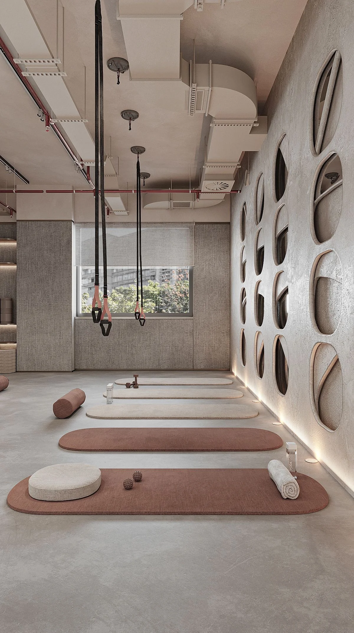 A yoga studio with mats, bolsters, and a foam roller on the floor. The studio has a large window with a view of buildings and trees outside. The wall on the right has a decorative design with circular cutouts, and there are hanging straps from the ce