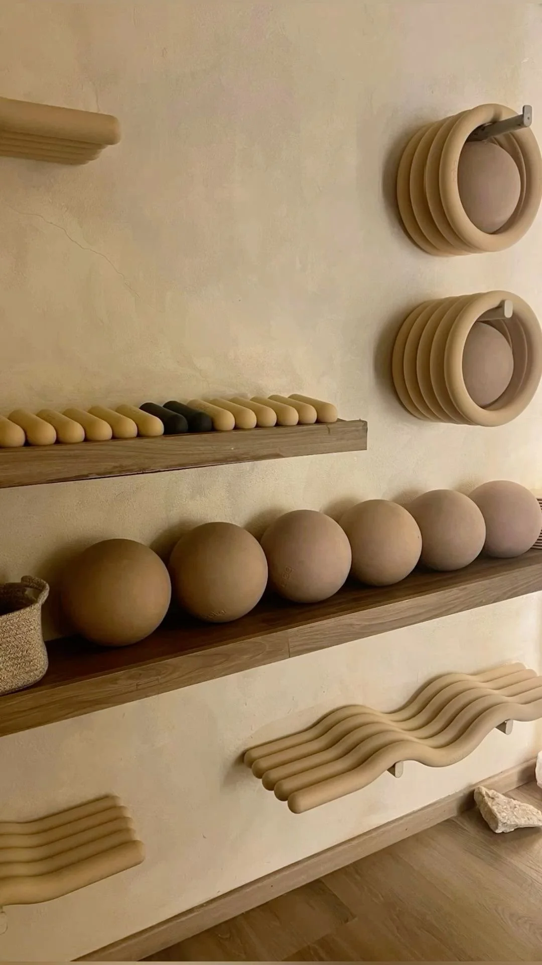 Display of wooden massage rollers, balls, and wave-shaped tools on wooden shelves on a beige wall.