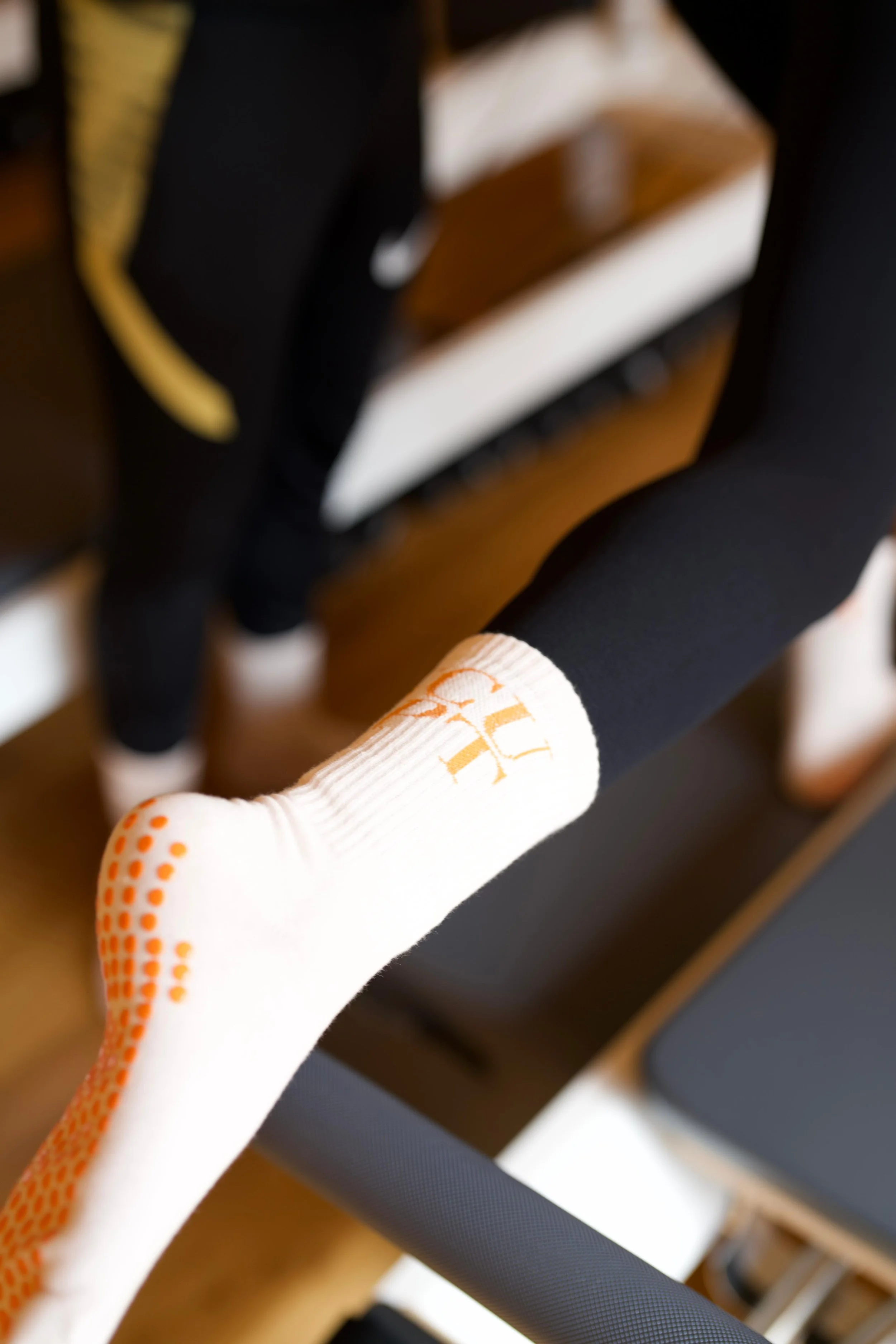 Close-up of a person wearing a white sock with orange dots and orange writing, and black pants with orange accents, standing near gym equipment.