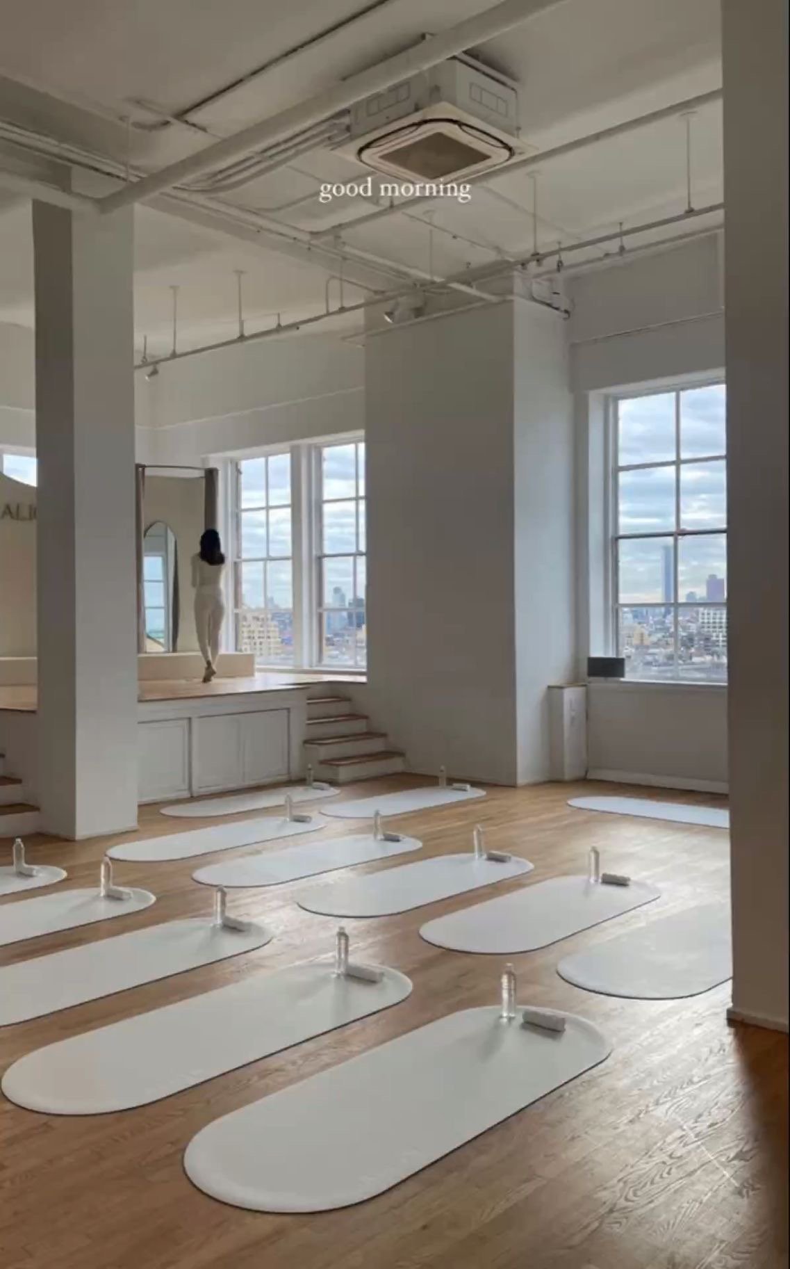 A yoga or fitness studio with white mats on wooden floors, resistance bands, and large windows overlooking a cityscape. The room has an industrial ceiling with an air conditioning unit and exposed pipes, with the words 'good morning' on the ceiling.