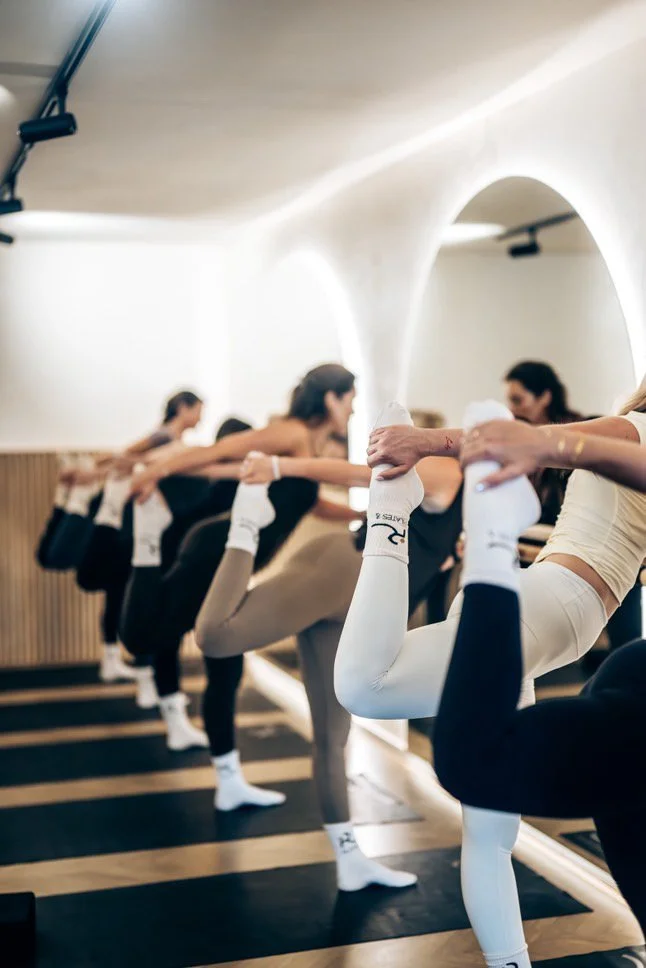 A group of women practicing yoga in a studio, balancing on one leg while holding their ankles with one hand, stretching their thighs and balancing with support from each other.