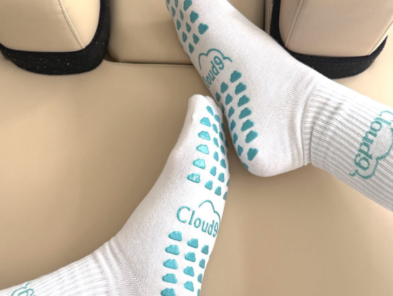 Person wearing white socks with blue cloud patterns and the word "Cloud" on the soles, resting on a beige car seat.