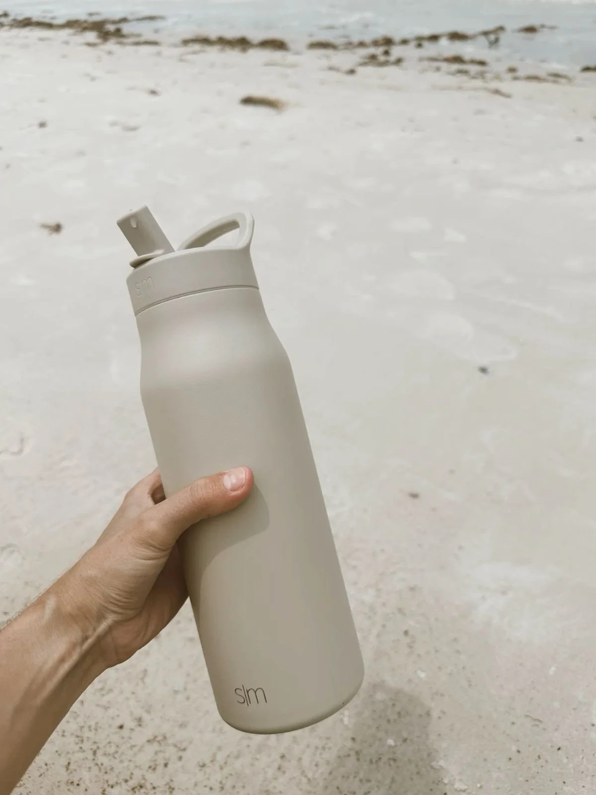 A person holding a beige reusable water bottle on a sandy beach, with the ocean in the background.