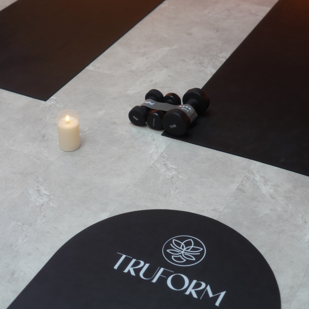 Yoga mat with the word 'TRUFORM' and a logo, black dumbbells on a black yoga mat, white candle on tiled floor.