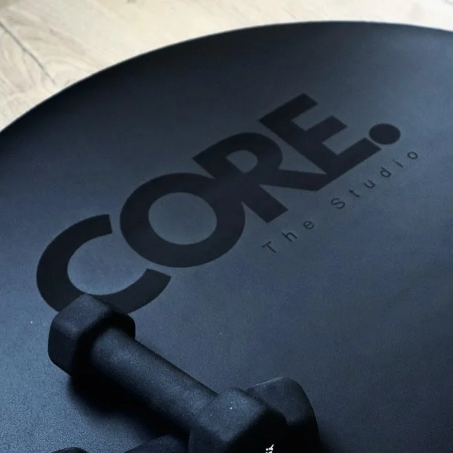 Close-up of a black exercise or yoga ball with the word "CORE" and "The Studio" printed on it, along with part of a black dumbbell resting on top.