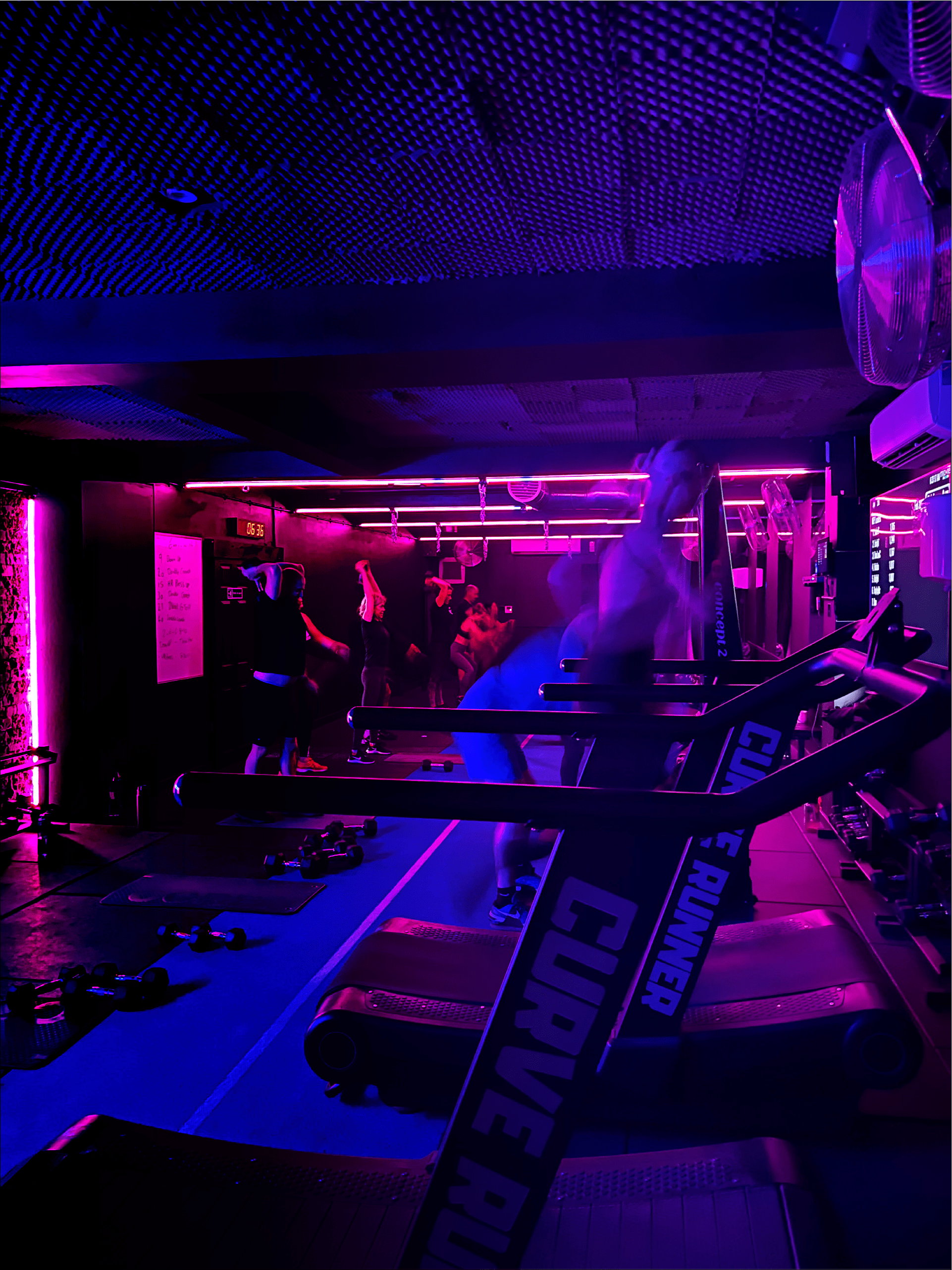 A dimly lit fitness studio with purple and pink neon lighting. People are exercising with various gym equipment, including treadmills and weights. A large sign reading 'STRENGTH' is visible in the foreground.