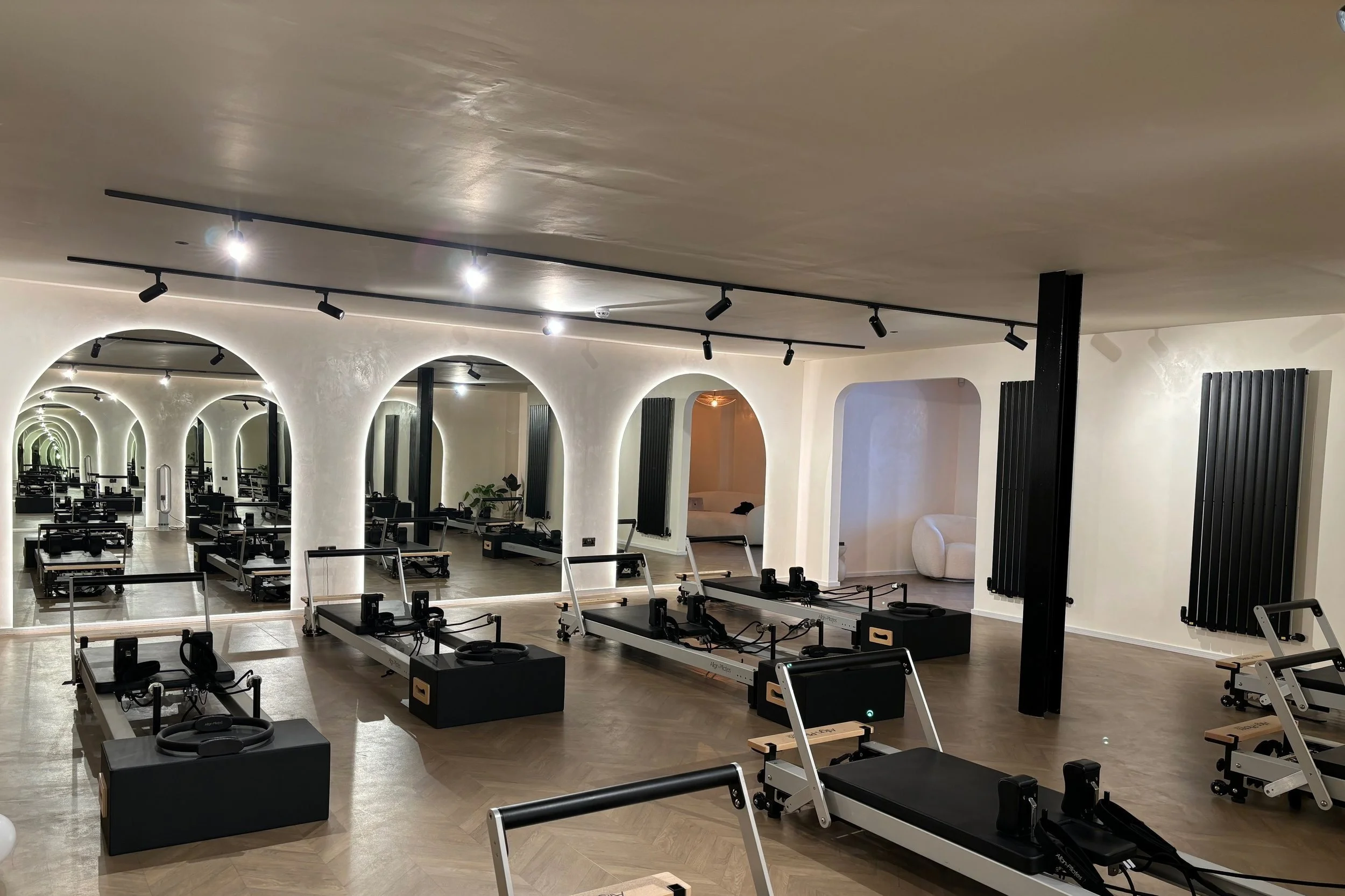 Fitness studio with reformer Pilates machines arranged in rows, mirrored walls, and modern lighting fixtures hanging from the ceiling.