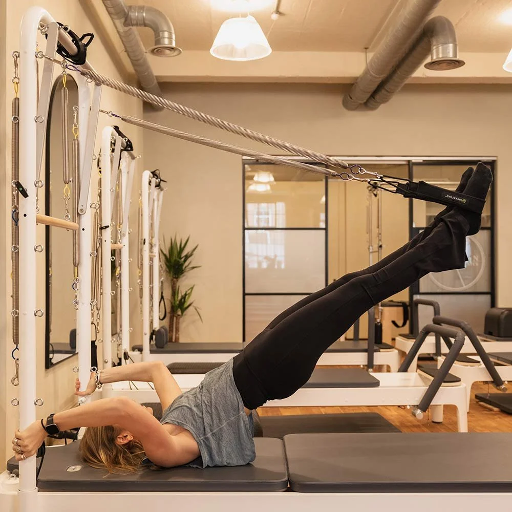 A woman is lying on a Pilates reformer machine, holding the handles with her legs extended upward, engaging in a workout at a gym.