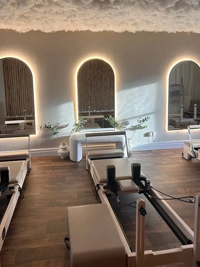 Spa room with three reformer Pilates machines, a small table, decorative plants, and three illuminated arched mirrors on the wall.