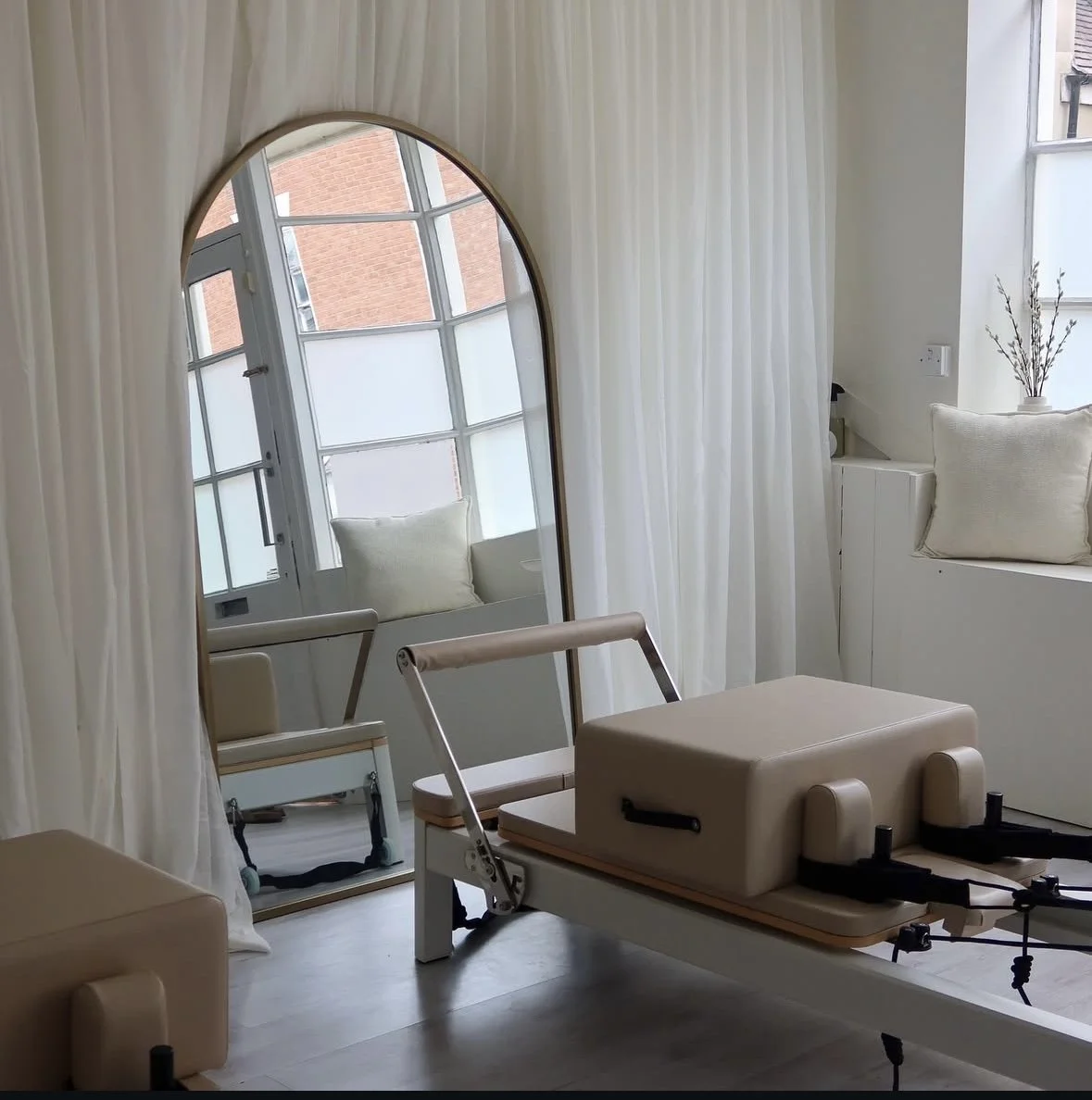 A room with a large arched mirror, a window seat with cushions, a potted plant, white curtains, and fitness equipment on a white treadmill with a cushioned platform.