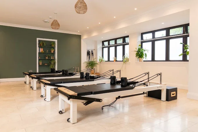 A Pilates studio with four reformer machines, large windows, and green plants on a white shelf and window ledge.