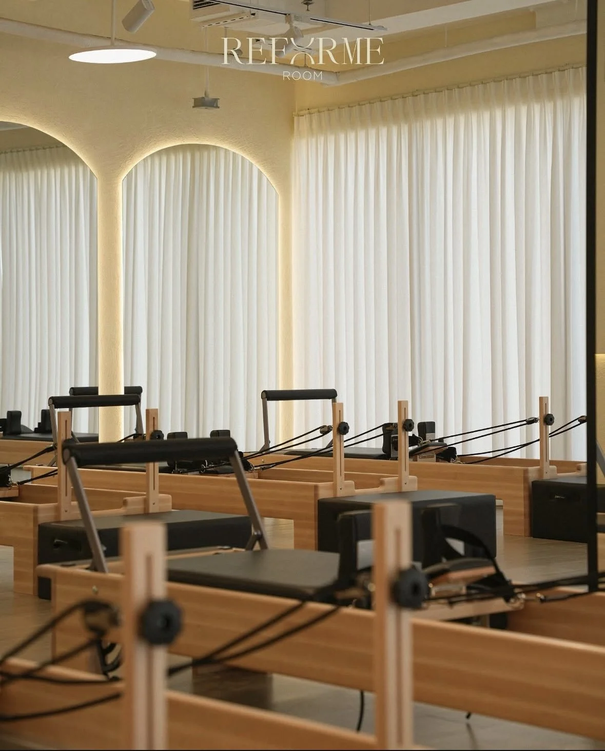 A Pilates studio with wooden reformer machines arranged in a row, large white curtains covering the windows, and soft ambient lighting.