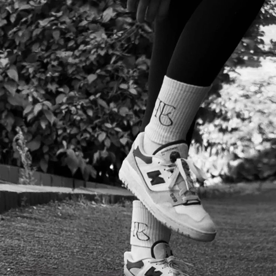 Close-up of a person's feet wearing Nike sneakers and socks with the word 'YES' written on them, standing on the edge of a paved path with foliage in the background.