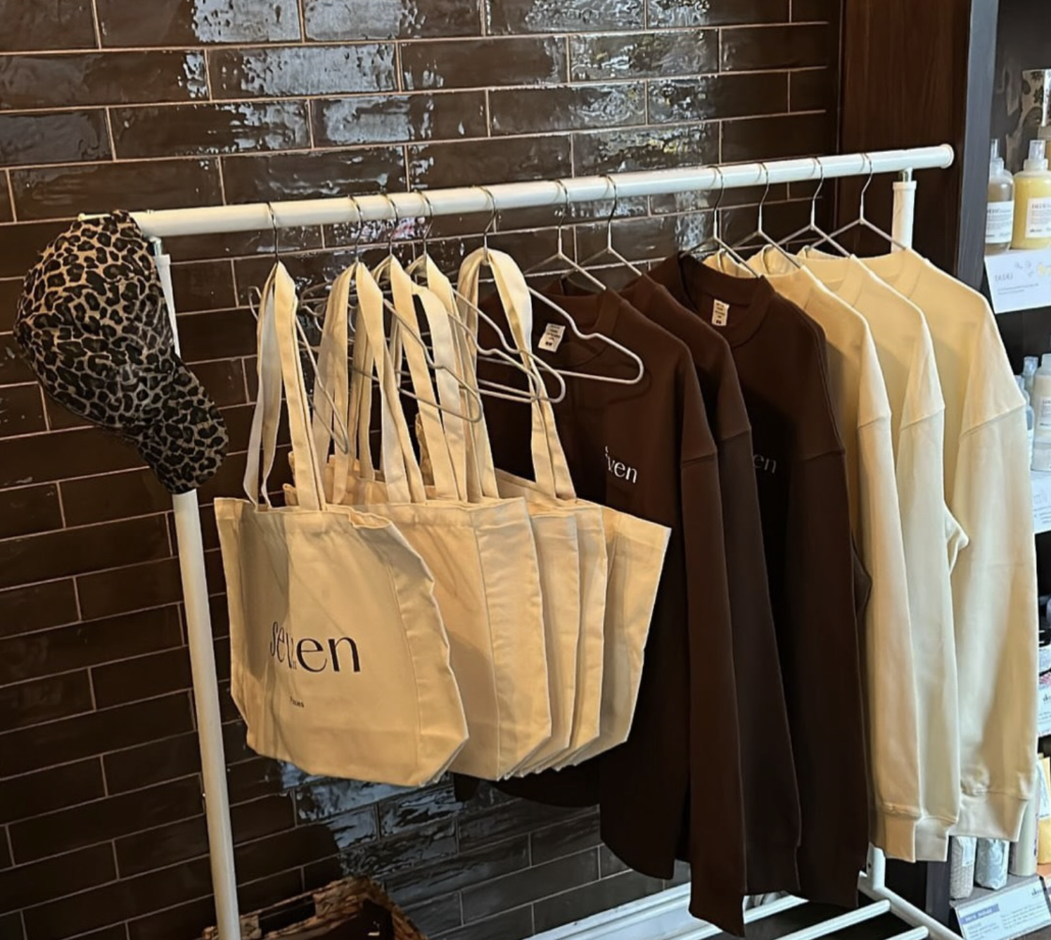 Clothing display on a rack, including cream tote bags with the word 'seven' on them, brown and beige sweatshirts, and a leopard print sleep cap, against a brown tiled wall with shelves holding bottles.