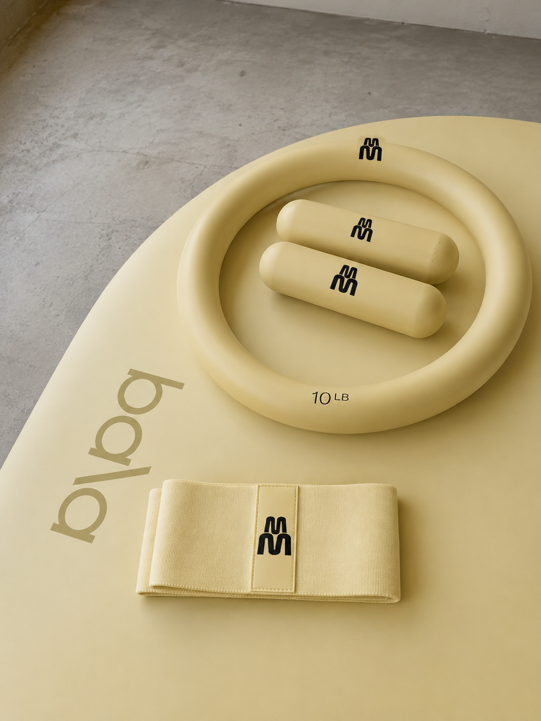Display of beige fitness accessories, including a resistance band labeled '10 LB', two cylindrical handles labeled 'S2', and a fabric workout belt, all arranged on a beige surface with the brand name 'body' partially visible.