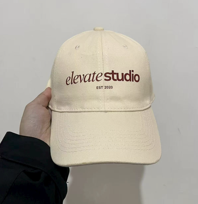 A beige baseball cap with the words "elevate studio" and "EST 2020" embroidered on the front, held by a person's hand against a plain wall.