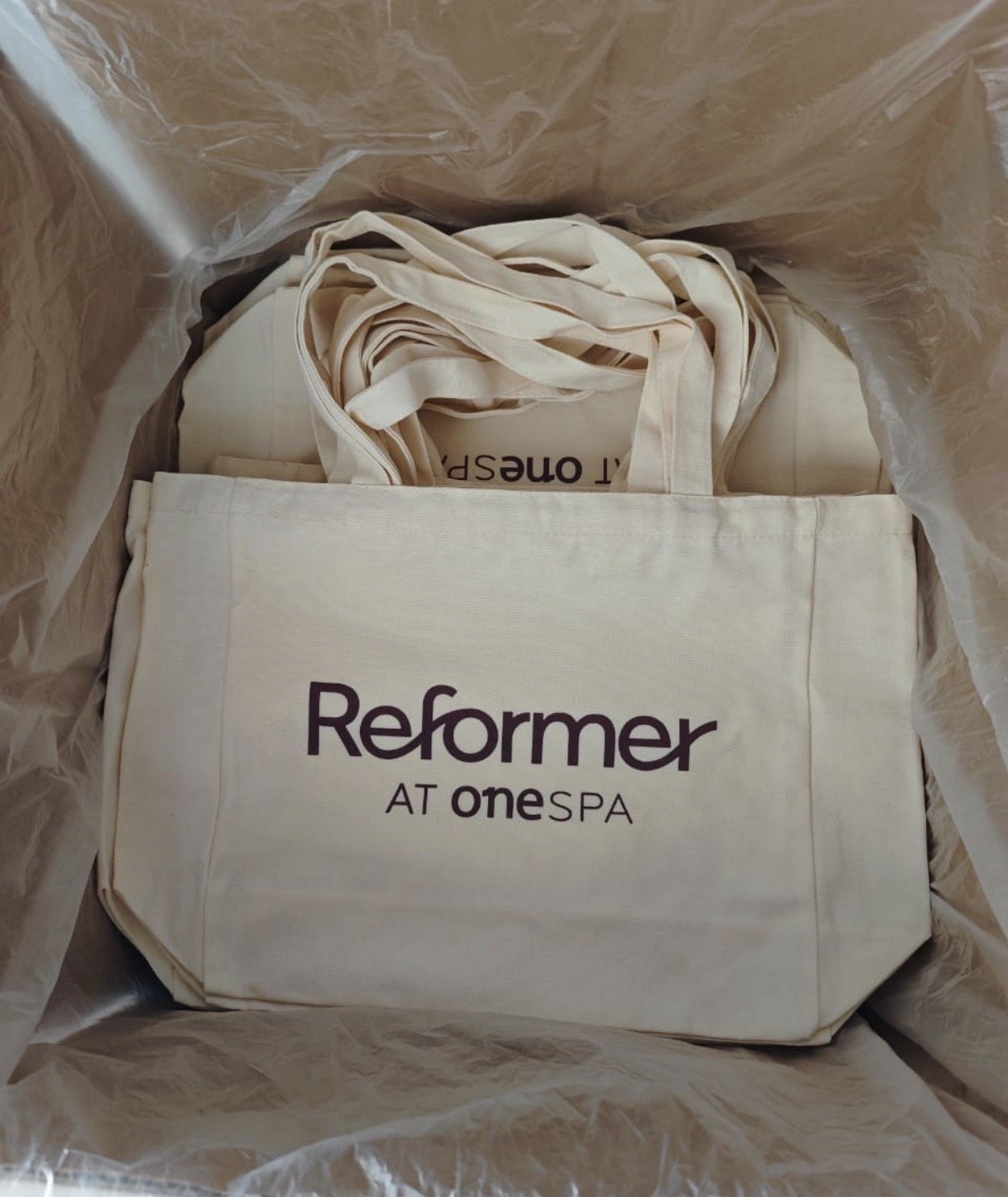 A reusable beige tote bag with the text 'Reformer AT onespa' printed on it, placed inside a plastic bag.