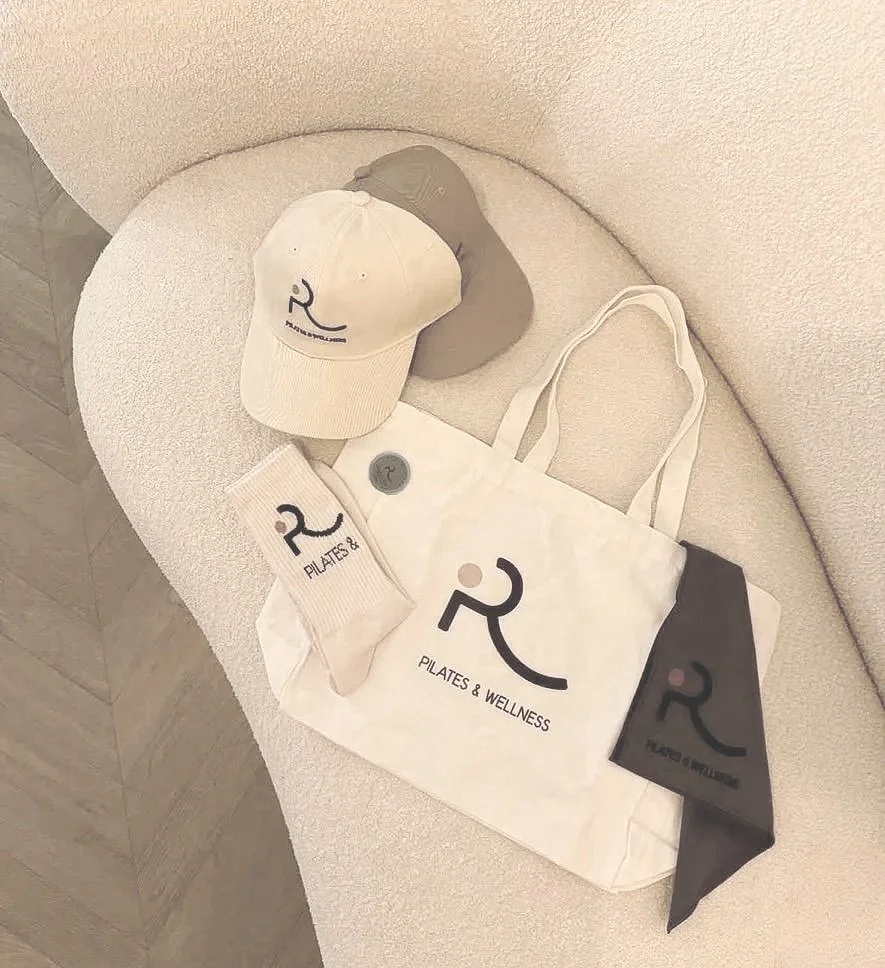 A beige cap, white and black tote bags, socks, and a small round pin, all branded with the Pilates & Wellness logo, are arranged on a textured beige seat.