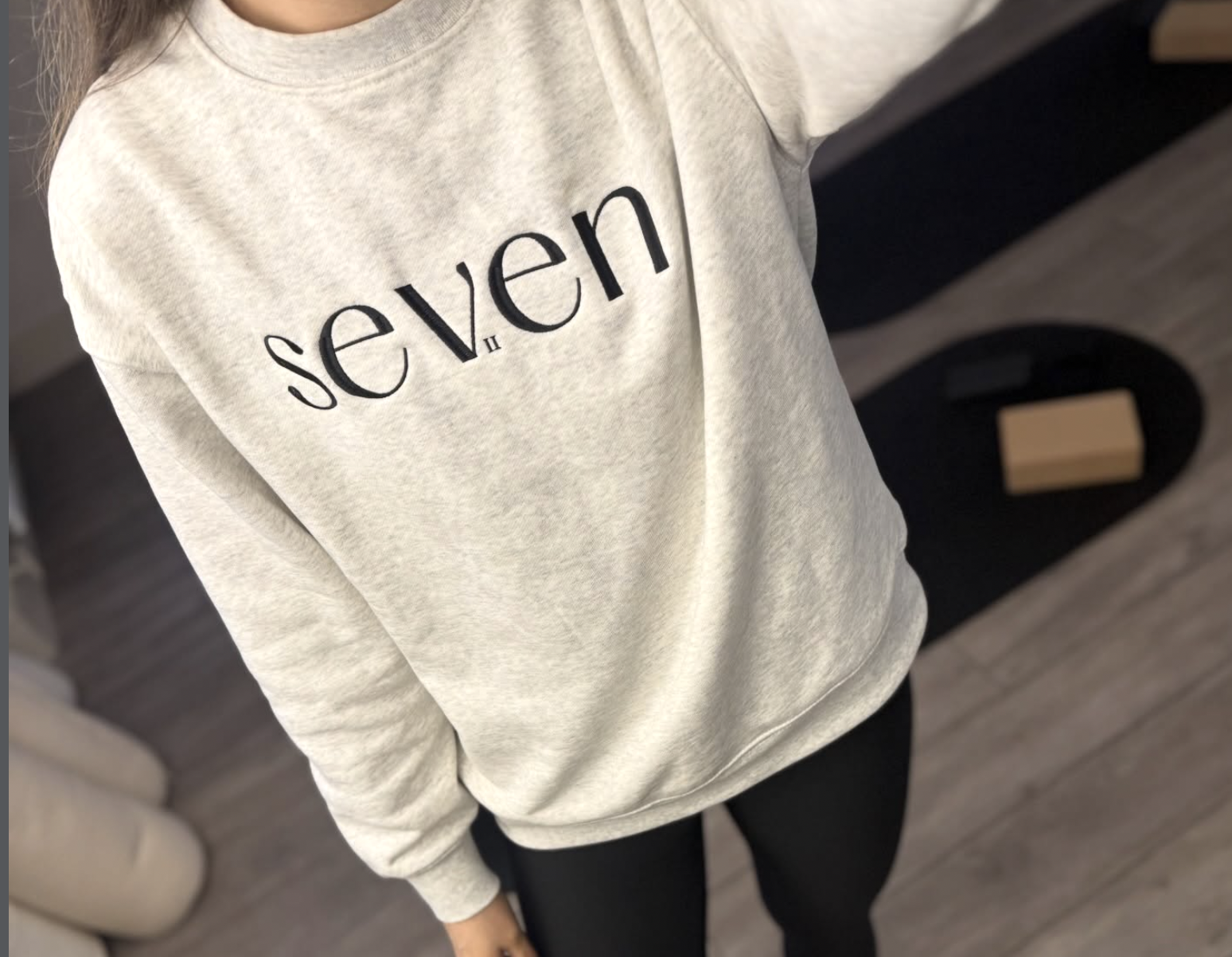 A person wearing a light gray sweatshirt with the word 'seven' printed on it, standing indoors on a wooden floor.