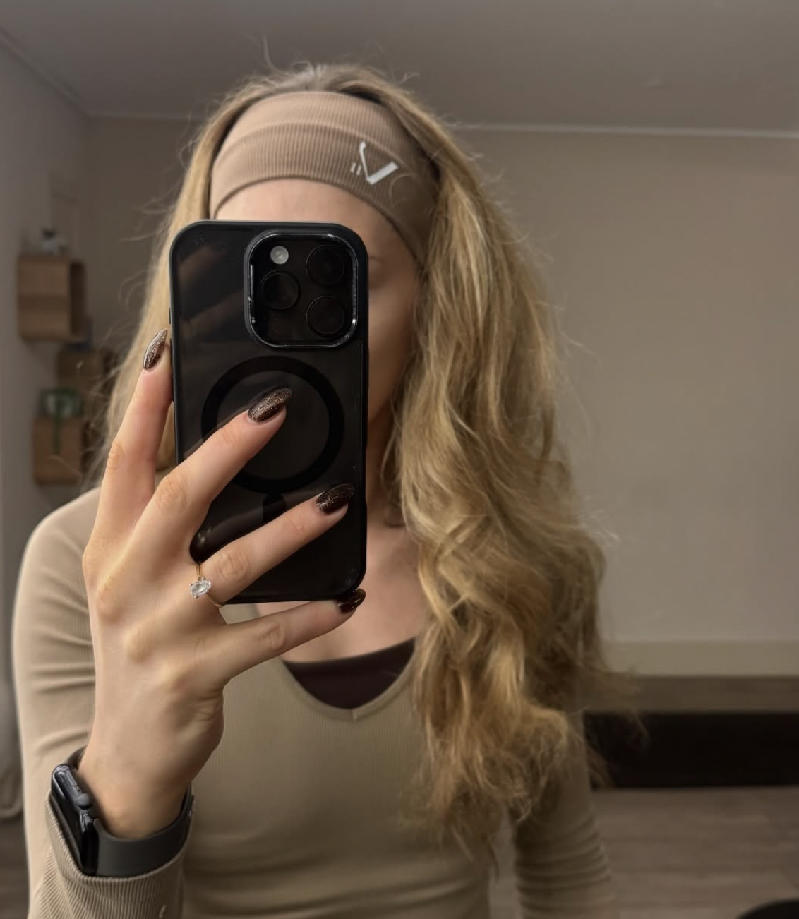 A woman with long, curly blonde hair takes a mirror selfie with her smartphone, wearing a beige headband and beige long-sleeve top.