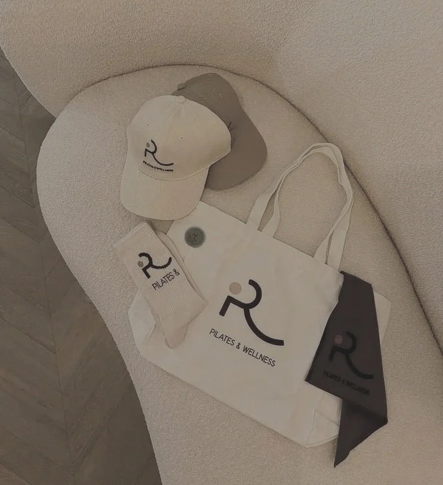 A white tote bag, a beige baseball cap, a pair of beige socks, and a black cloth face mask, all with a logo featuring a stylized person doing Pilates and the words "Pilates & Wellness," are arranged on a light-colored sofa.