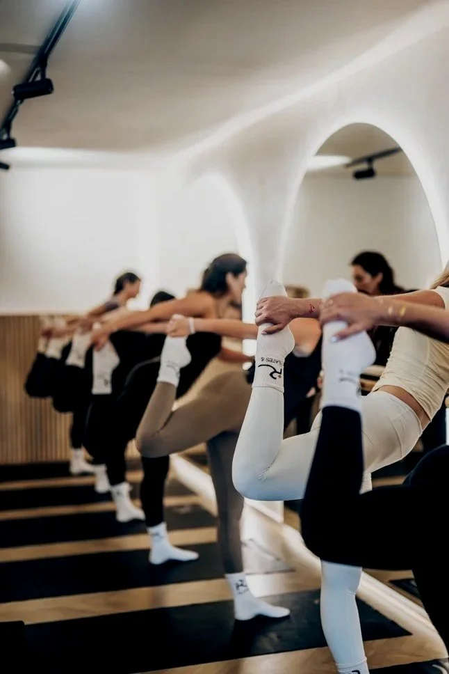 Women participating in a yoga class, practicing a balancing pose while holding onto a barre for support, in a calm studio with modern architecture.