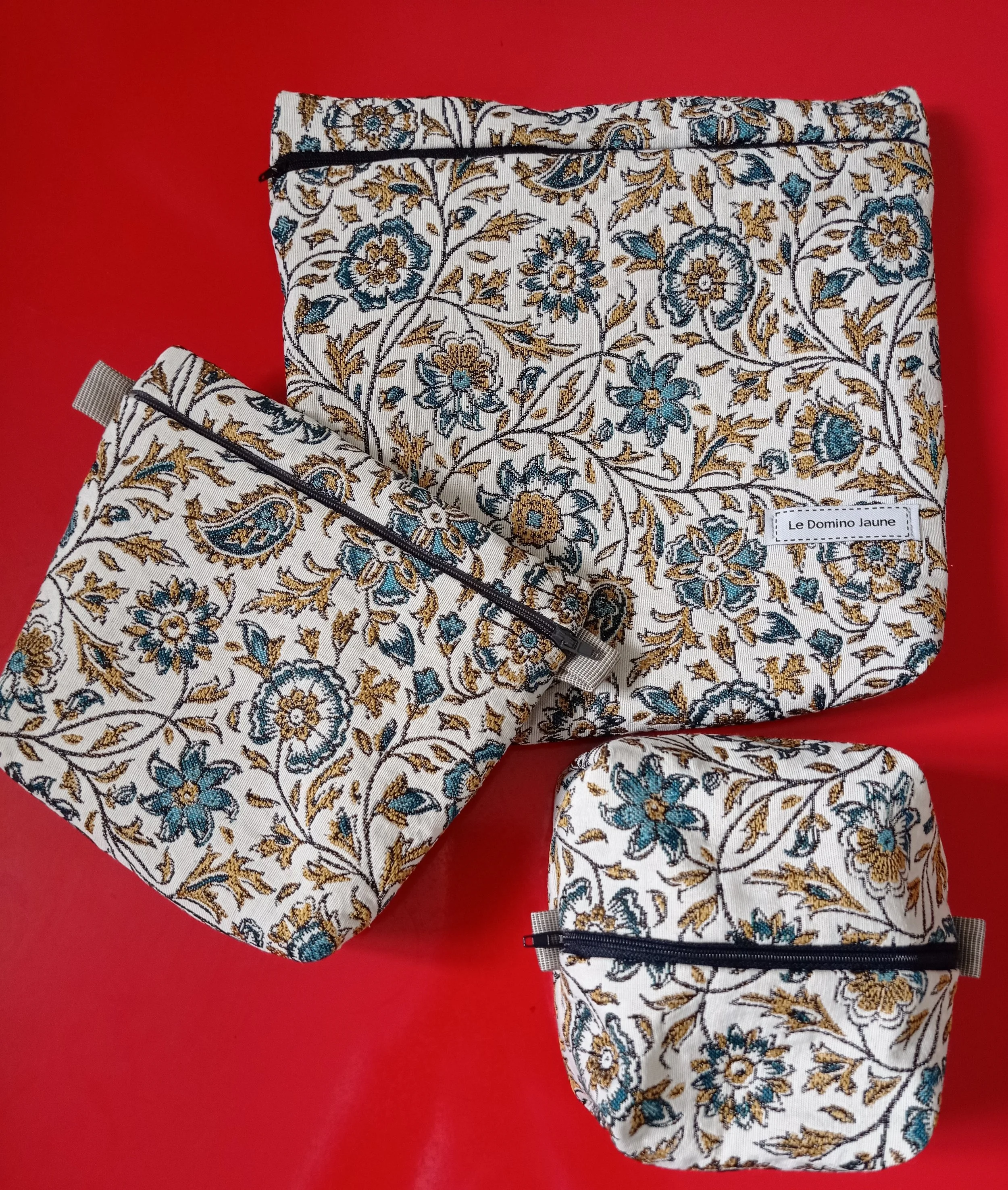 Ensemble de sacs de rangement en tissu à motifs floraux dorés, bleus et noirs, avec zips, posé sur un fond rouge.