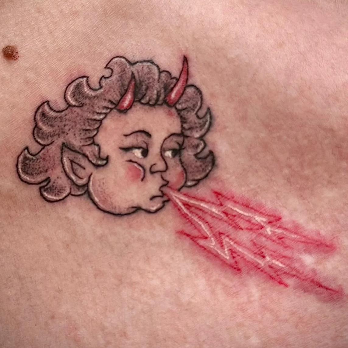 Color tattoo of a horned cherub face breathing red lightning bolts, whimsical illustrative style