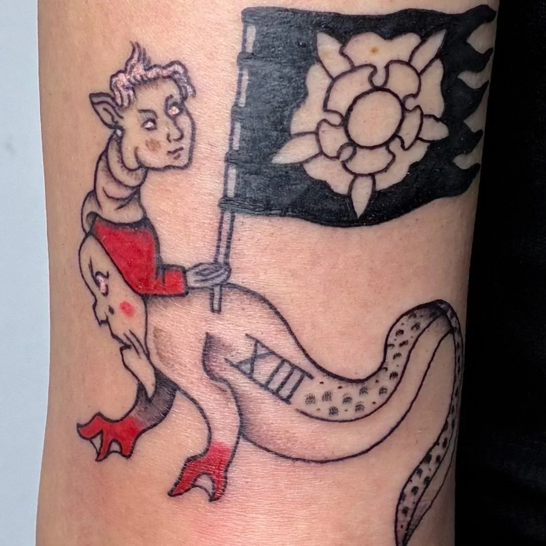 Color tattoo of a devil holding a tarot card flag with a heraldic crest, riding a snake-tailed creature with roman numerals