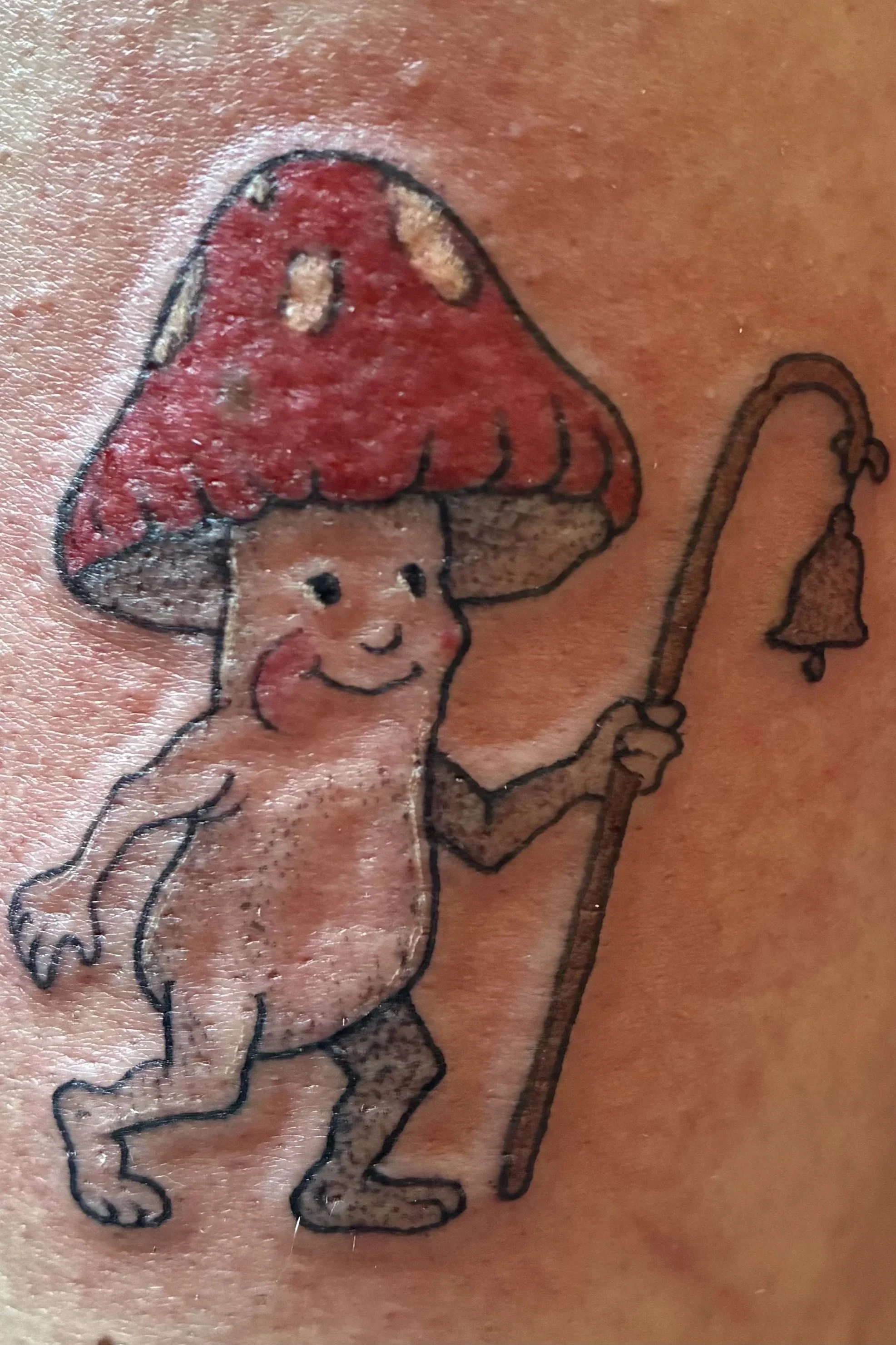 Color tattoo of a smiling mushroom character holding a staff with a bell, whimsical fantasy design