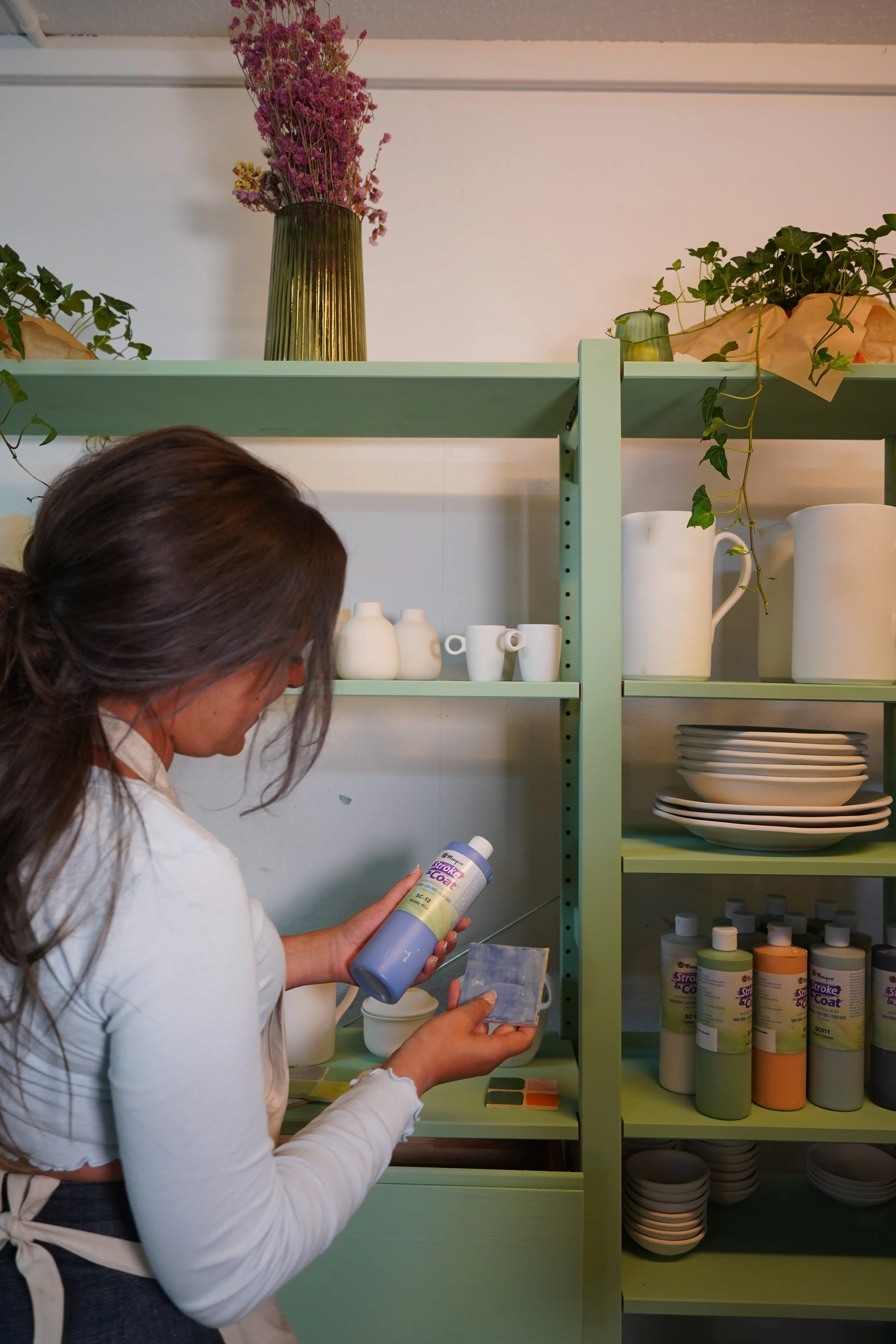 Woman in white shirt and apron holding a bottle of coating spray and a sample swatch in a kitchen or storage room with shelves of ceramics, plants, and decorative bottles.