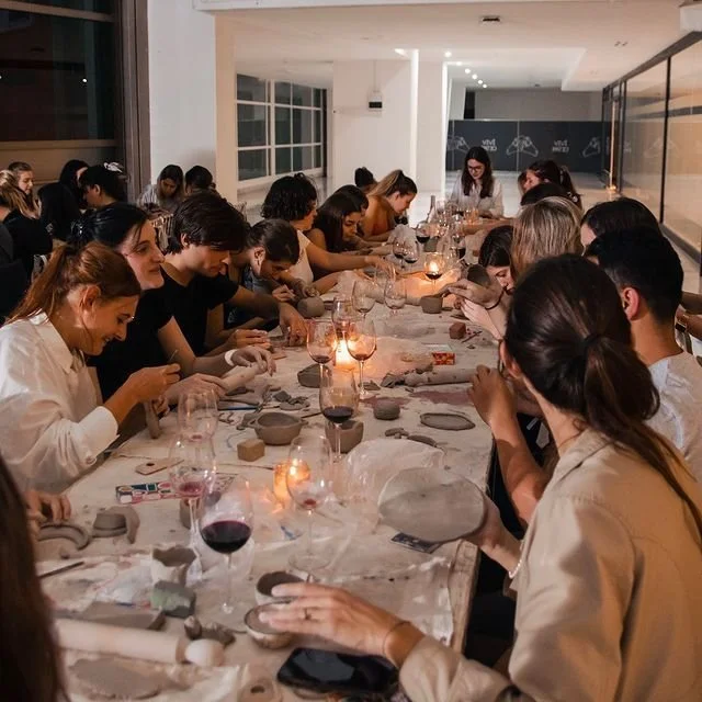 Group of people sitting around a long table, partaking in a pottery or ceramic painting activity, in a well-lit modern room.