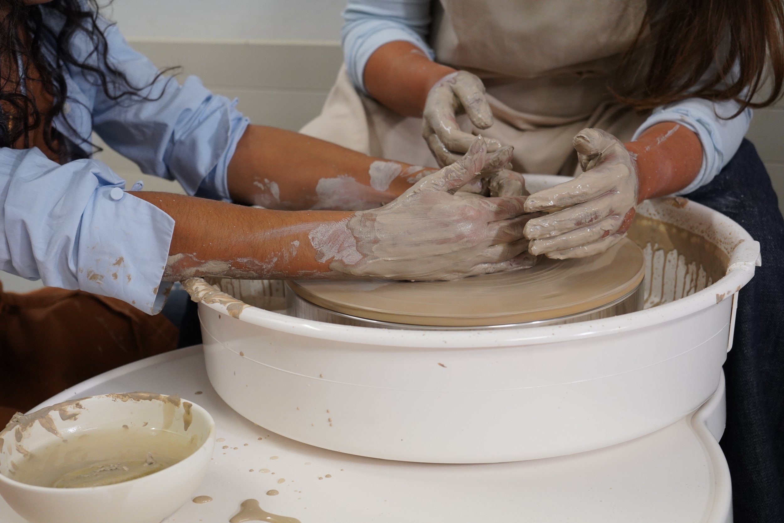People making pottery on a pottery wheel, with their hands covered in clay, in a studio setting.