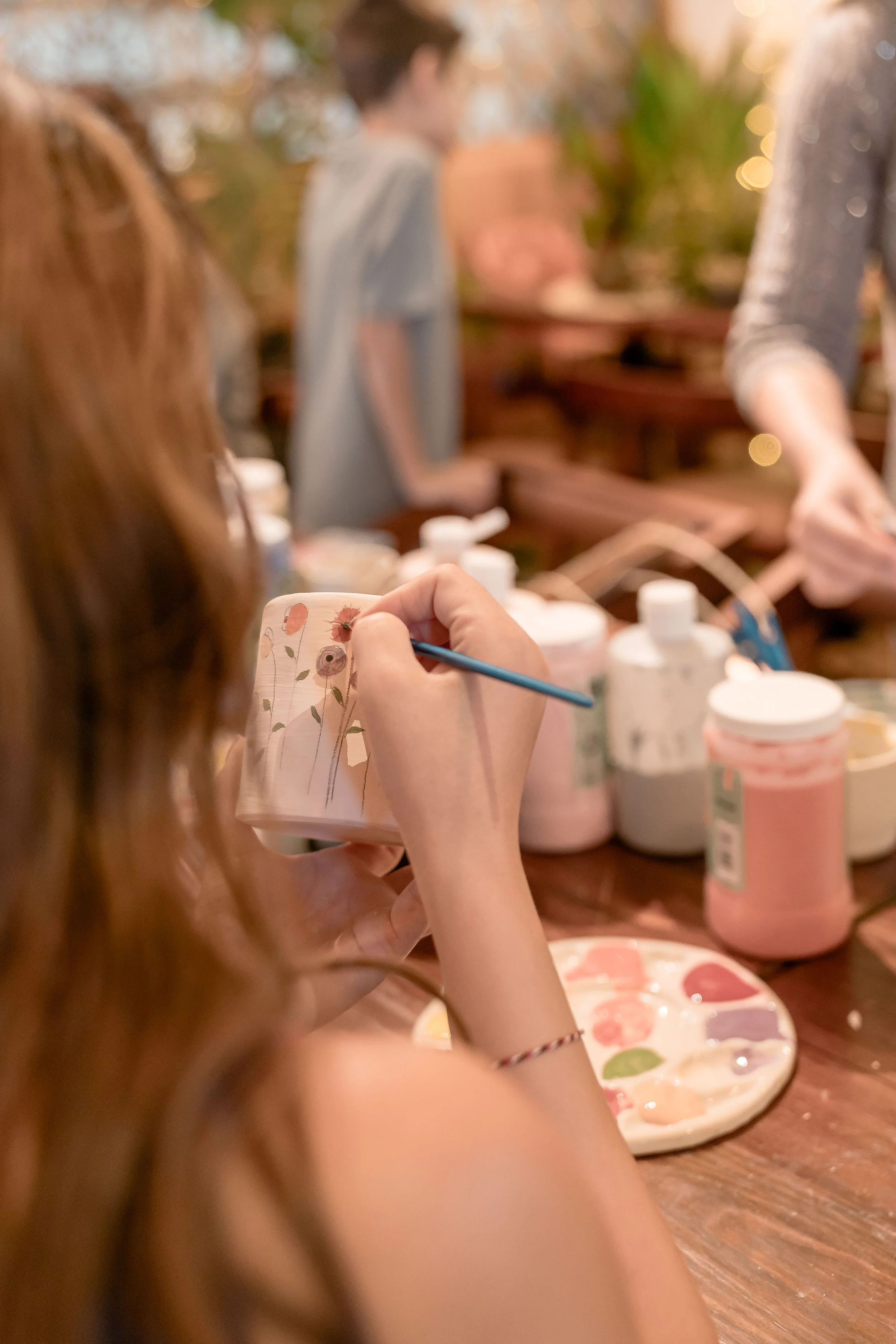 Close-up of a person painting on a ceramic mug with floral design, surrounded by paint bottles, and other people in the blurred background.